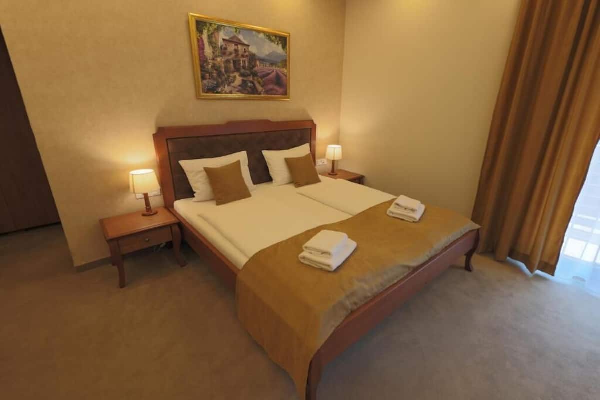 A hotel room with a large bed, two bedside tables with lamps, a painting on the wall, and a window with curtains.