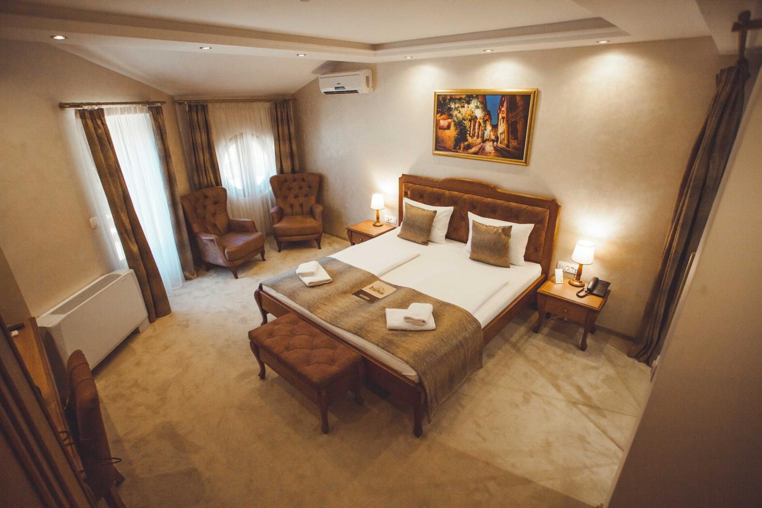 A hotel room with a large bed, two bedside tables with lamps, a chair, a bench, and a painting on the wall.