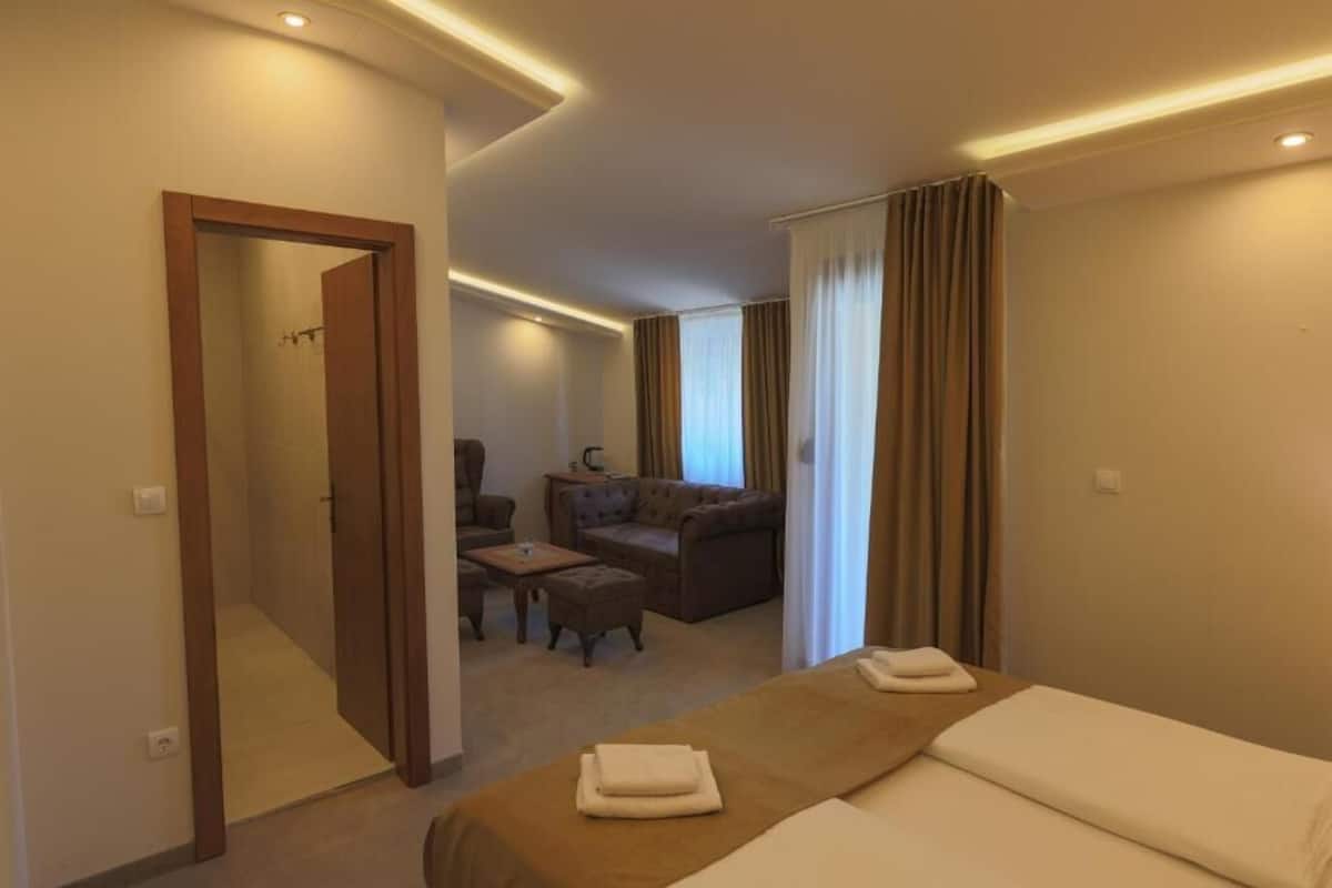 A hotel room with a bed, a sofa, a chair, and a small table. There is a door leading to a bathroom.