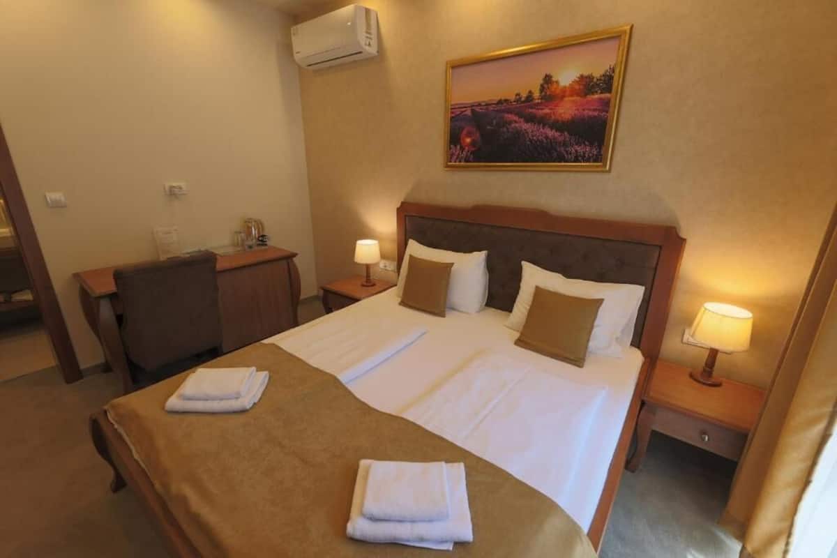 A hotel room with a double bed, two bedside tables with lamps, a desk with a chair, and an air conditioner on the wall.