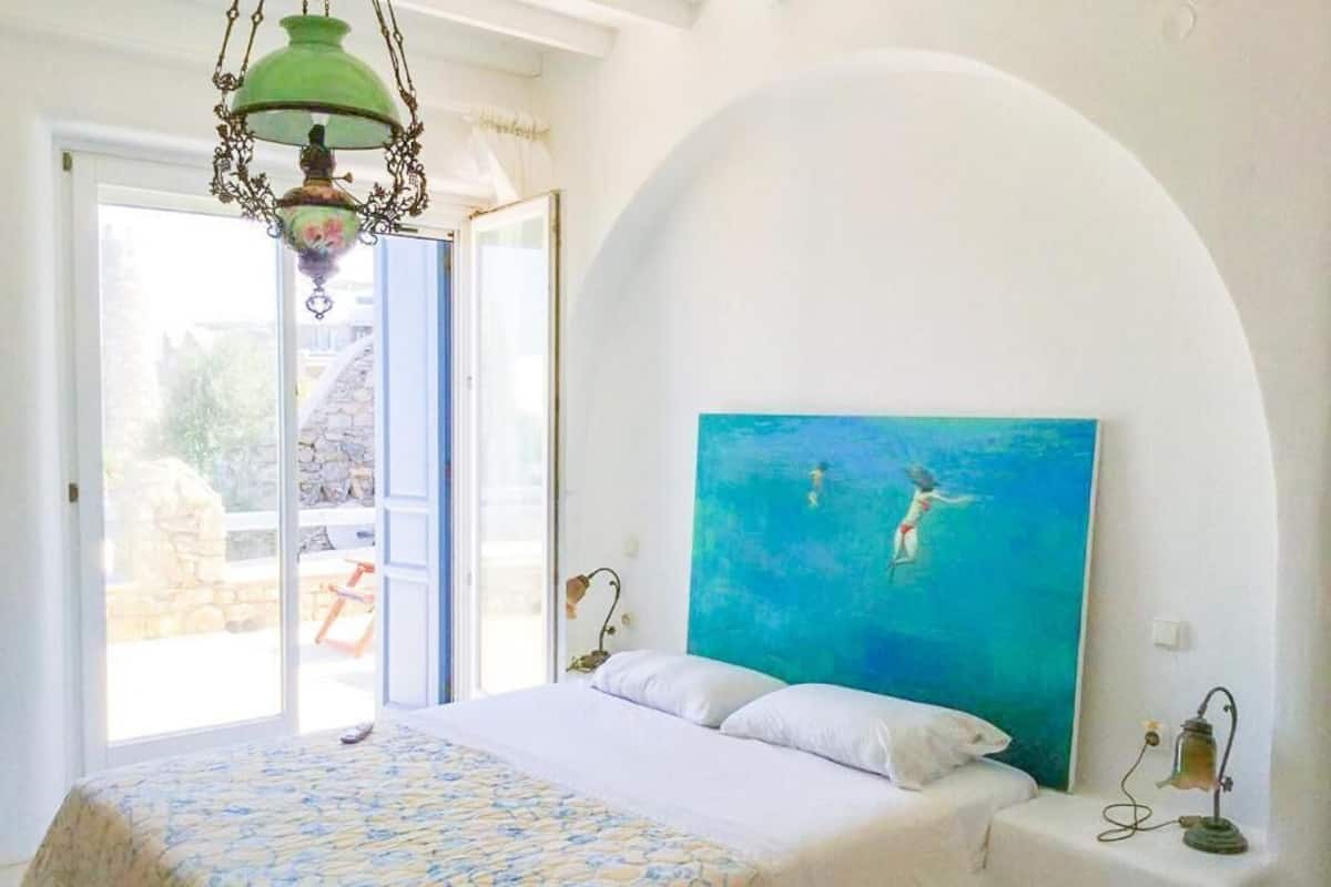 A bedroom with a bed, a painting of swimmers, a chandelier, and a view of the outside.