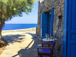 A stone building with blue shutters, a table with chairs, and a view of the sea. - Poseidon Secret Blue Pools (Mykonos)