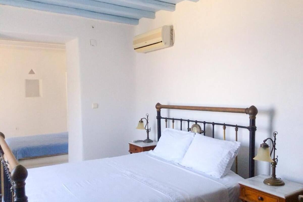 A bedroom with a wooden bed, white linens, two bedside tables with lamps, and a view of the sea through an open door.
