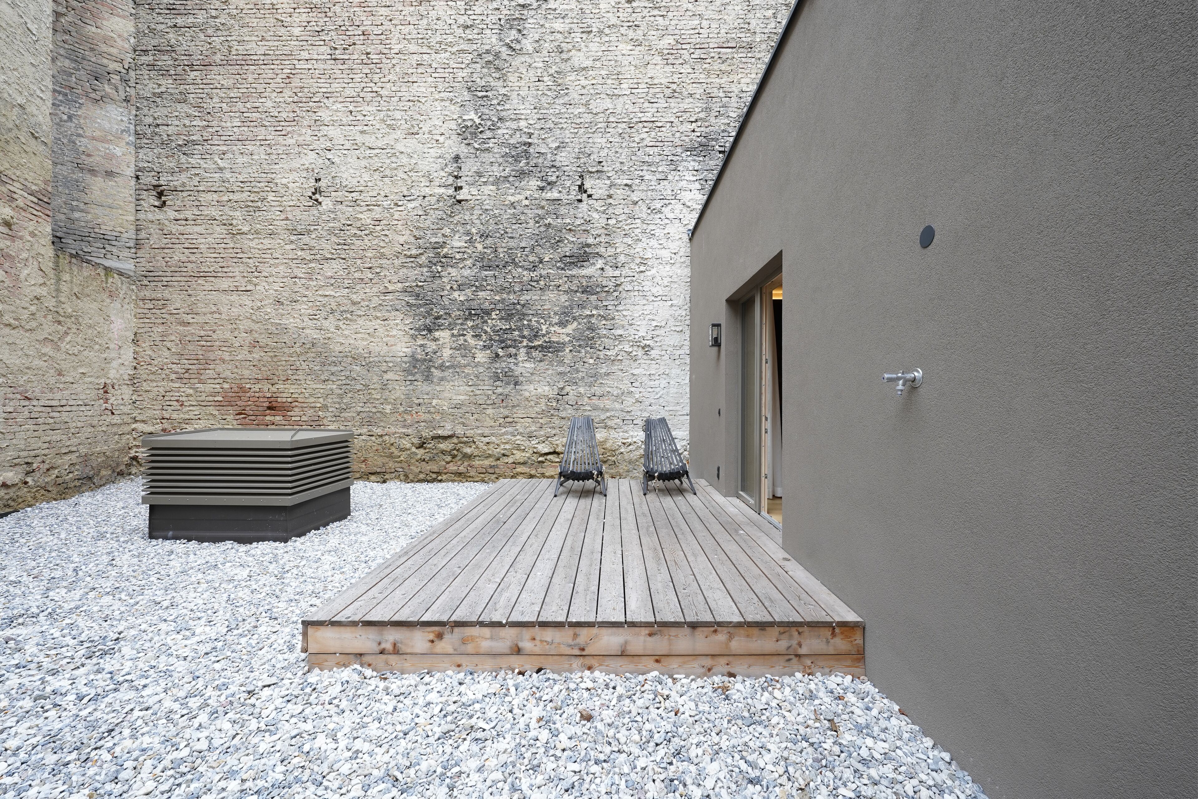 Une terrasse en bois avec deux chaises en métal, une pile de tapis et un bâtiment doté d’un mur texturé et d’une porte.