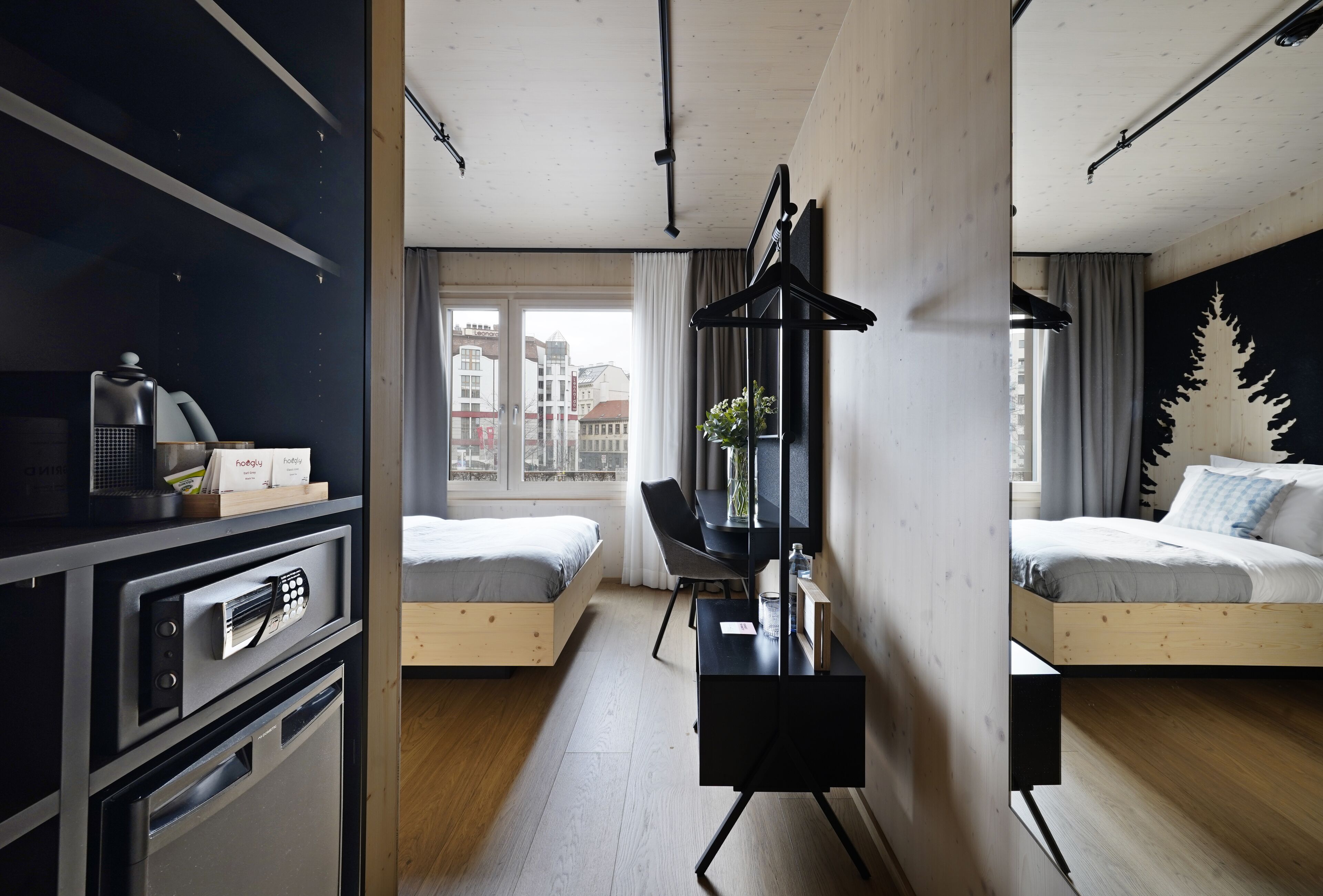 Une chambre d’hôtel moderne avec un lit en bois, un bureau avec une chaise, un porte-manteau noir suspendu et une grande fenêtre donnant sur la ville.
