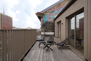 Una terraza de madera con dos sillones de descanso y una mesa, un mural en la pared contigua y vistas de la ciudad.