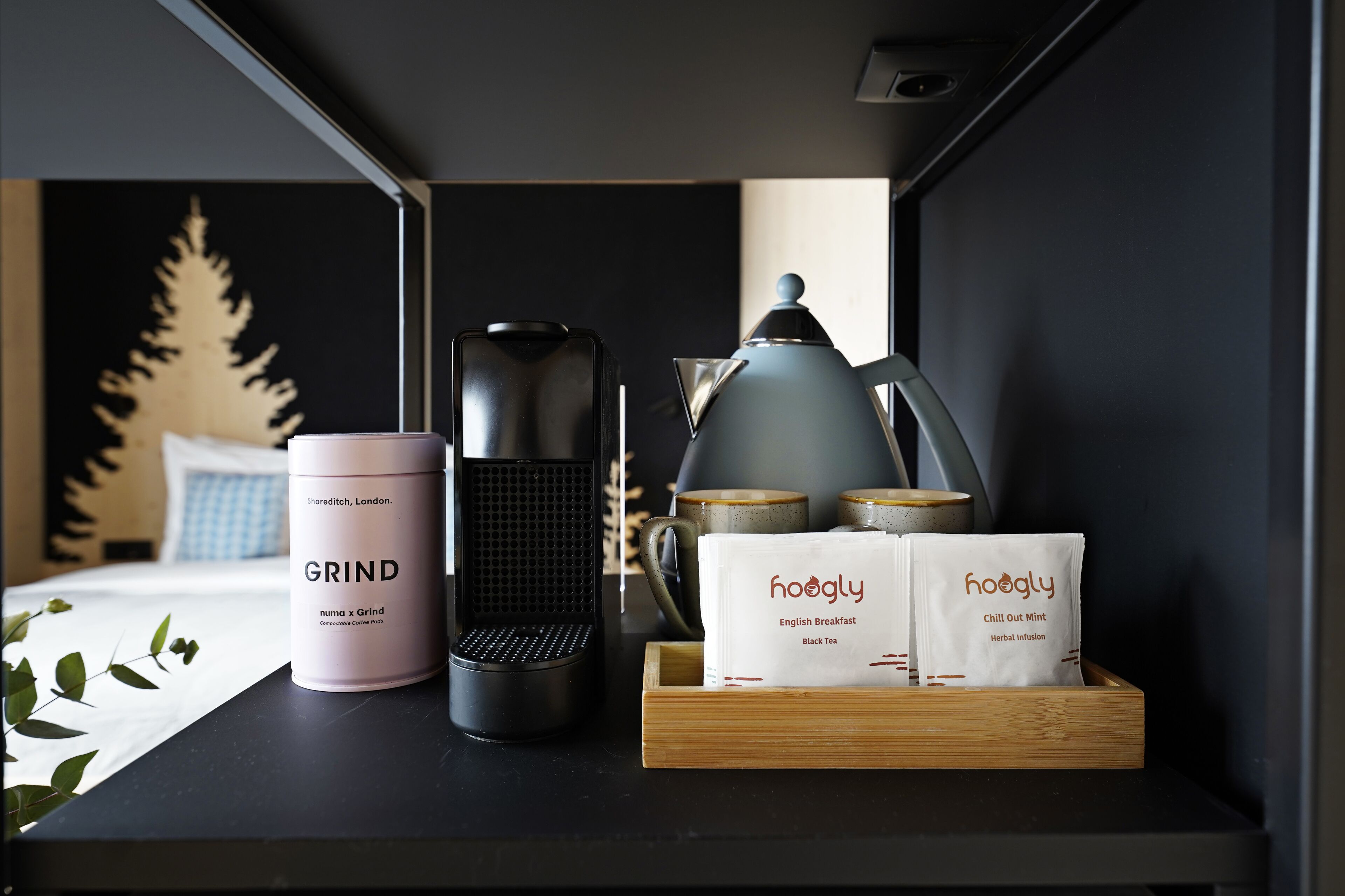 Uma estação de café com um moedor, uma chaleira e dois pacotes de infusão herbal 'hoogly' sobre uma bandeja de madeira.