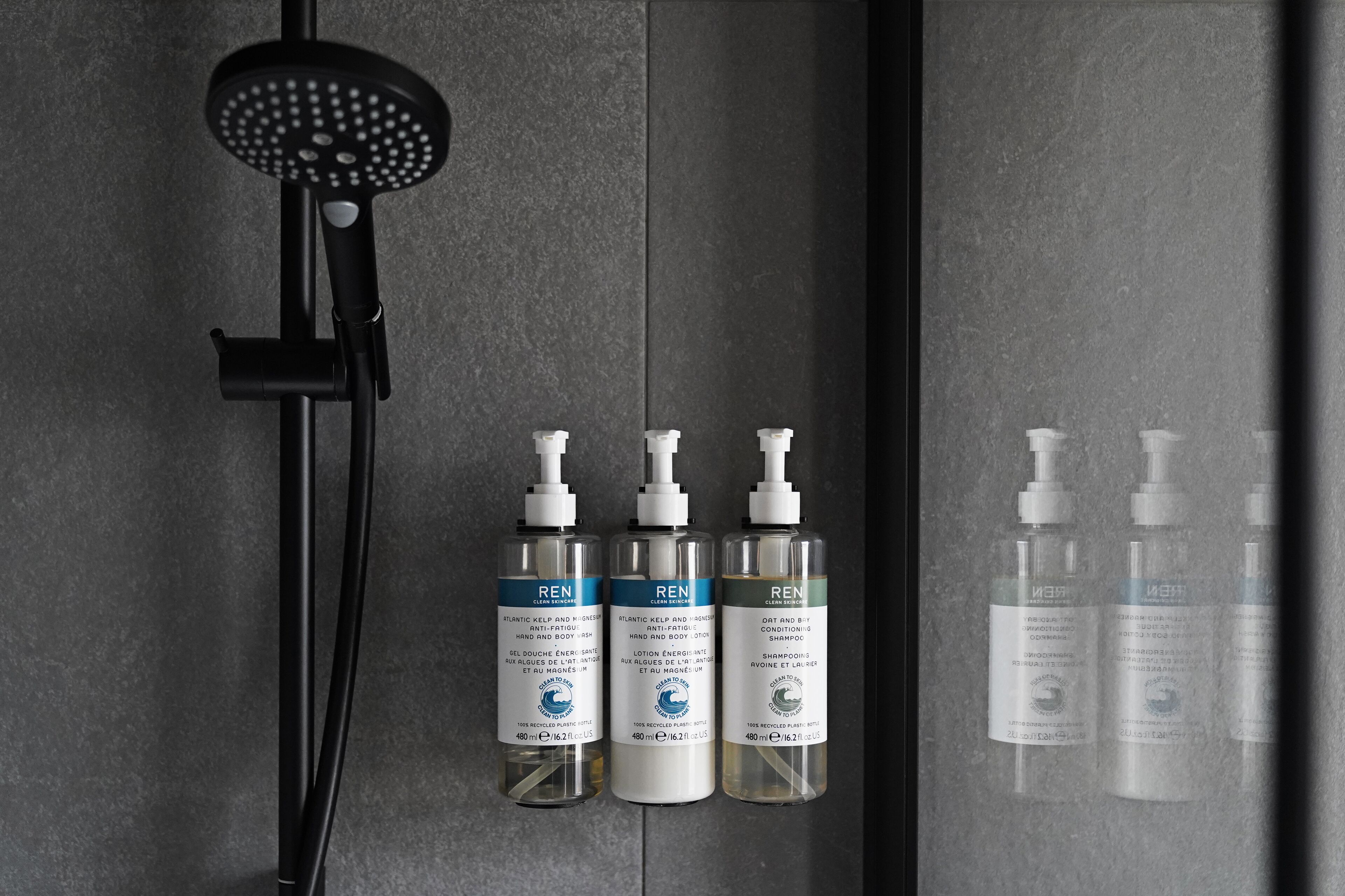 A shower area with a black handheld showerhead and three bottles of REN body wash and shampoo.