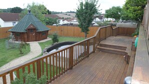 Une terrasse en bois offrant une vue sur un jardin, avec notamment un petit chalet en bois et un étang.