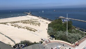 A coastal beach area with a lighthouse, sandy beach, and a promenade with people walking. - 80 m from the beach, everything in view - Anchorage vacation apartment (Rostock-Warnemünde)