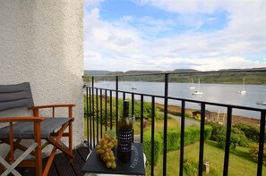 A balcony with a table holding a bottle of wine and a bunch of grapes, overlooking a lake with boats.