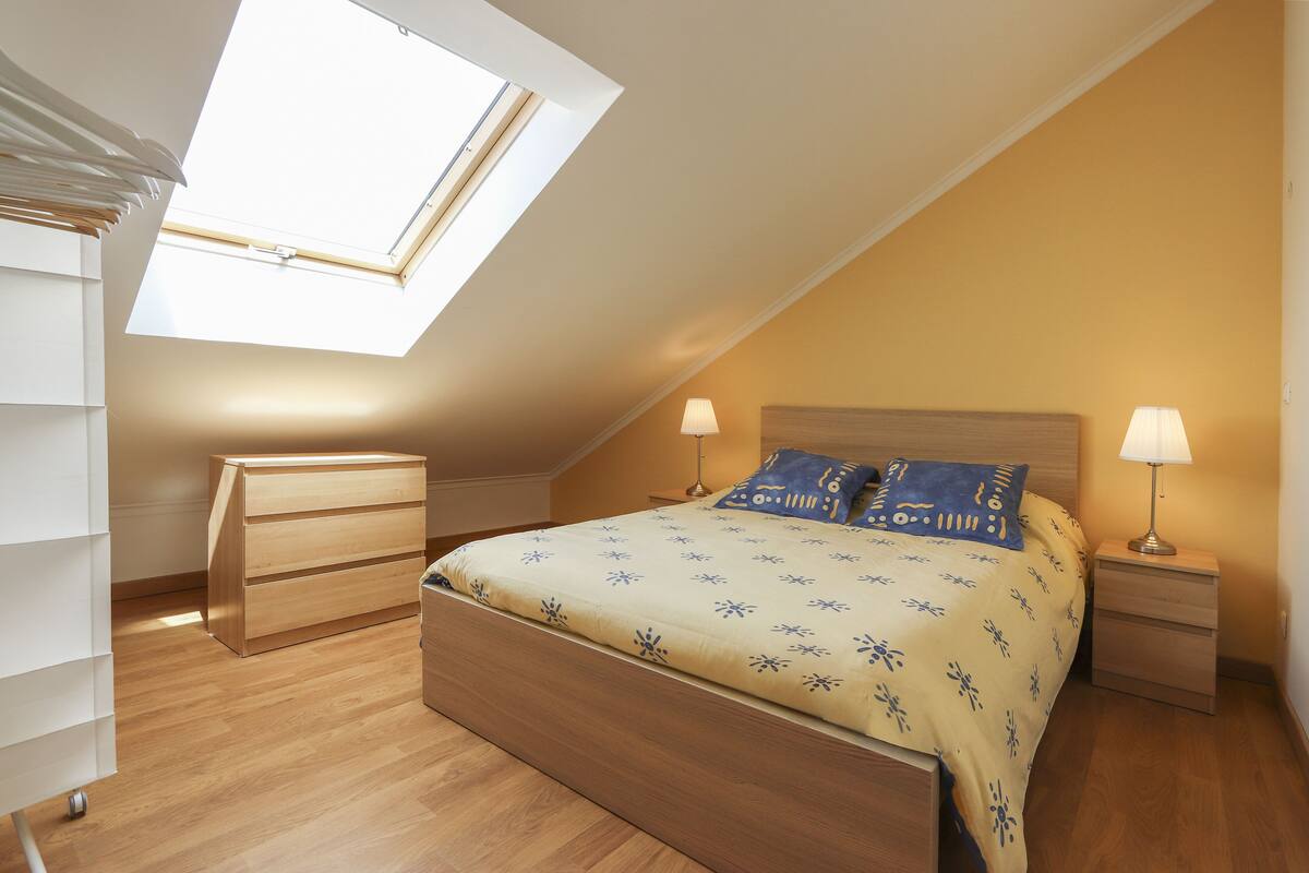 A bedroom with a skylight, a bed with patterned bedding, two bedside tables with lamps, and a wooden dresser.