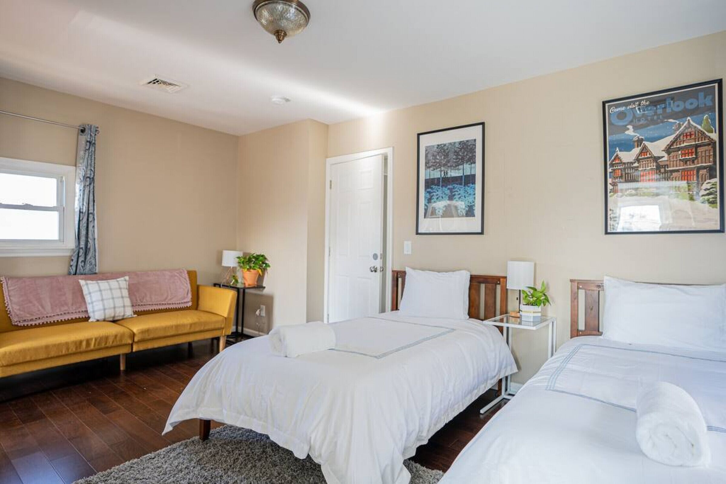 A hotel room with two beds, a sofa, a window with curtains, a door, and a wall with a framed picture.