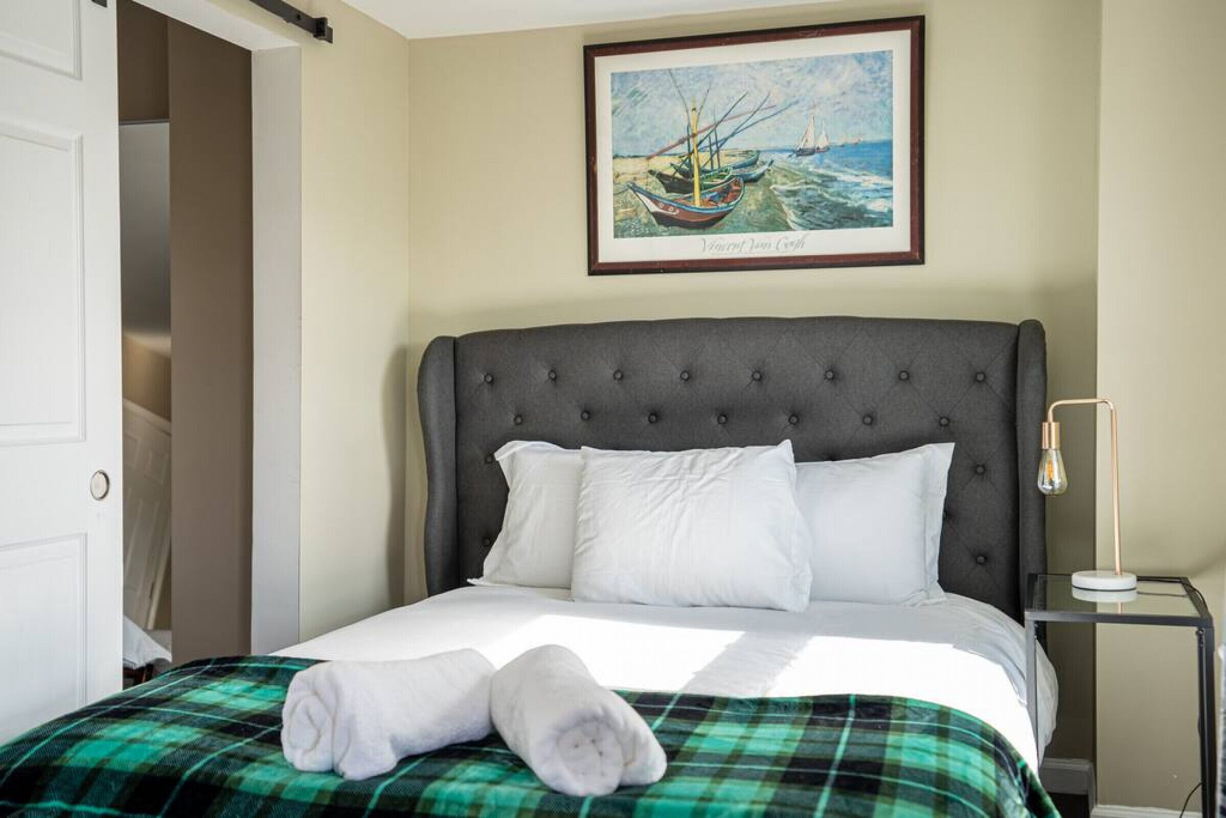 A neatly made bed with white linens and a plaid blanket, a framed painting of a boat on the wall, and a bedside table with a lamp.