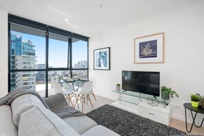 Un salon moderne avec un canapé, une table basse en verre, un téléviseur à écran plat et des œuvres d’art encadrées accrochées au mur.