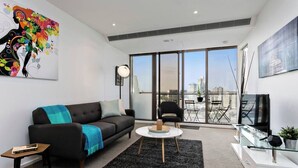 A modern living room with a sofa, coffee table, and TV stand, large windows with a city view, and artwork on the walls.