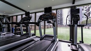 A gym with treadmills, ellipticals, and a view of a tennis court through large windows.