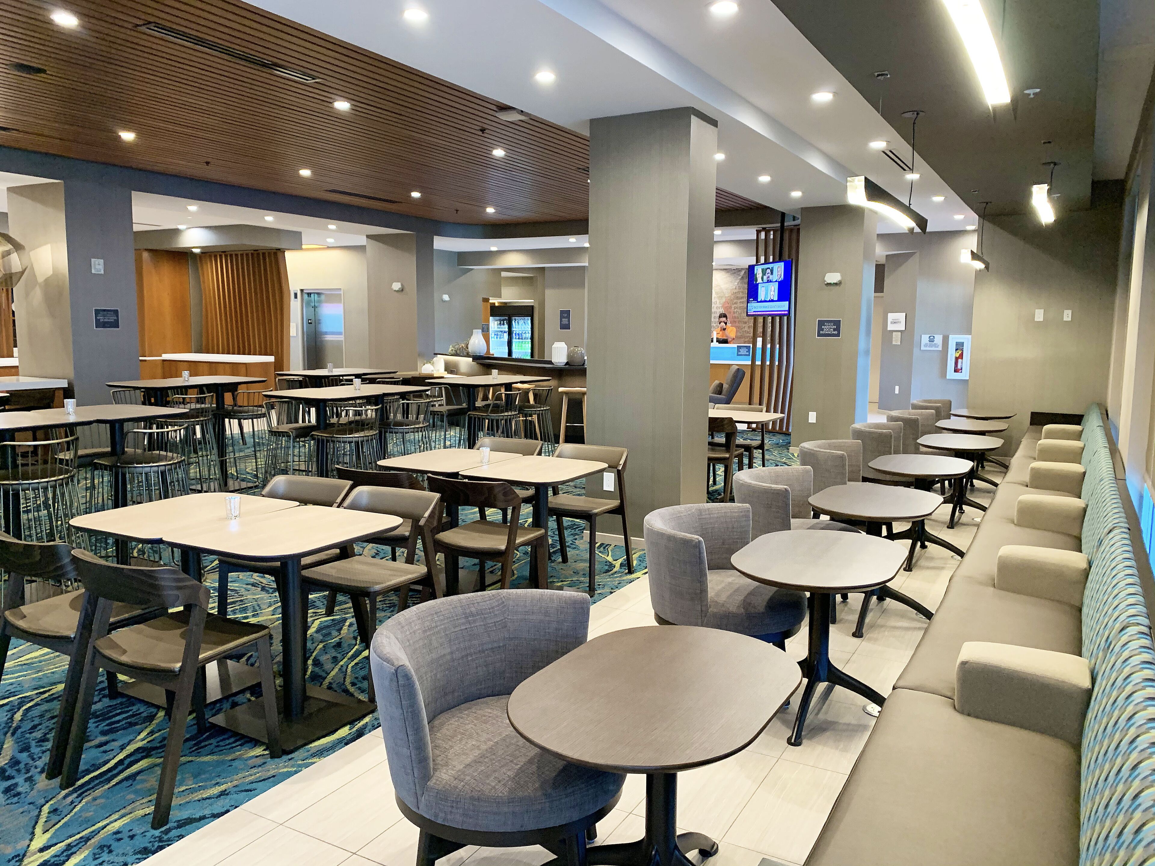 A hotel lobby with multiple seating areas, including tables and chairs, and a reception desk with a television.