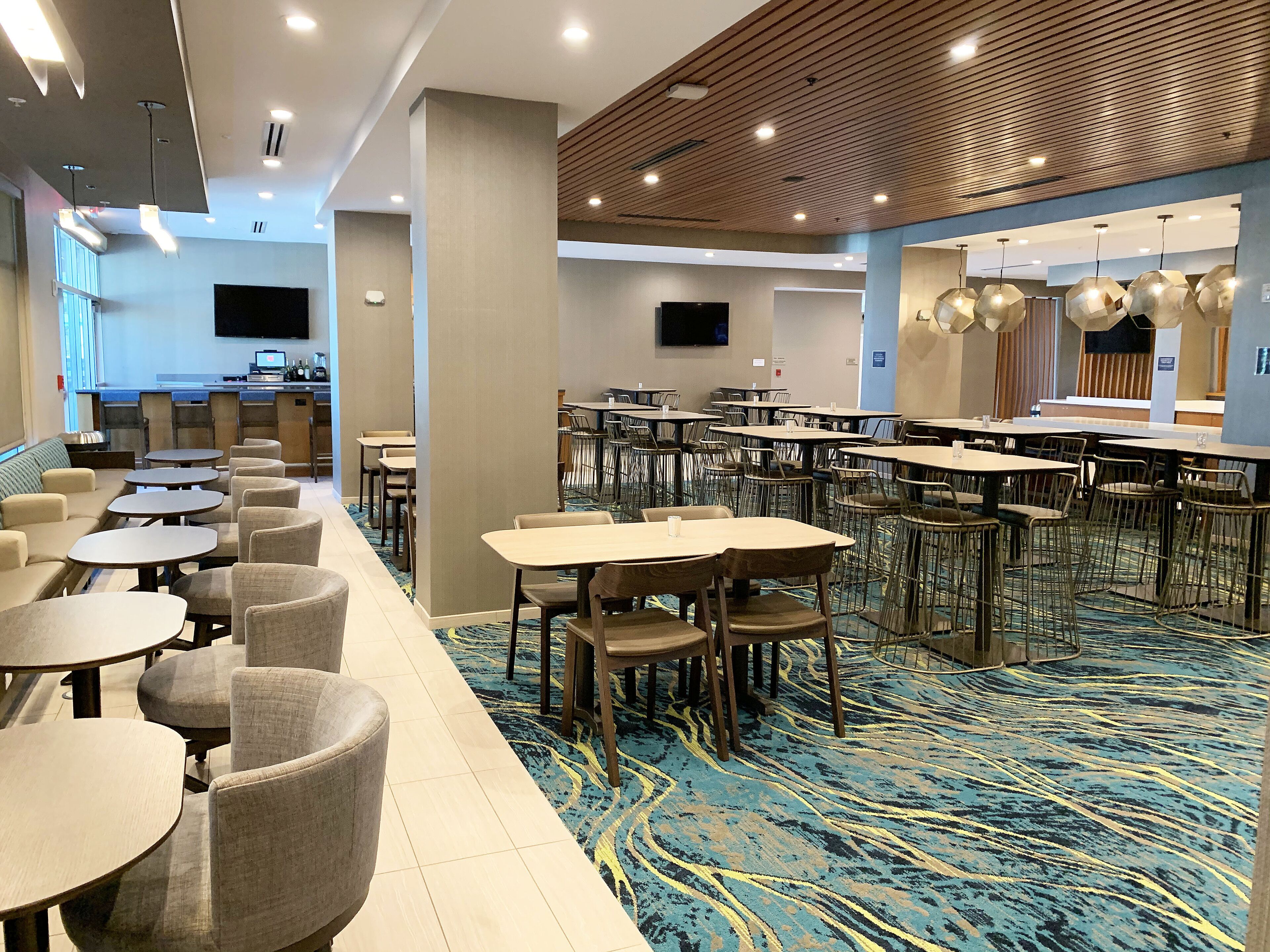Comedor de hotel con varias mesas y sillas, zona de cocina con televisor de pantalla plana y alfombra estampada.