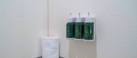 Un baño con una toalla blanca sobre un sujetabrazos plateado y tres botellas verdes de champú de árbol de té, acondicionador y jabón corporal de limón y salvia.