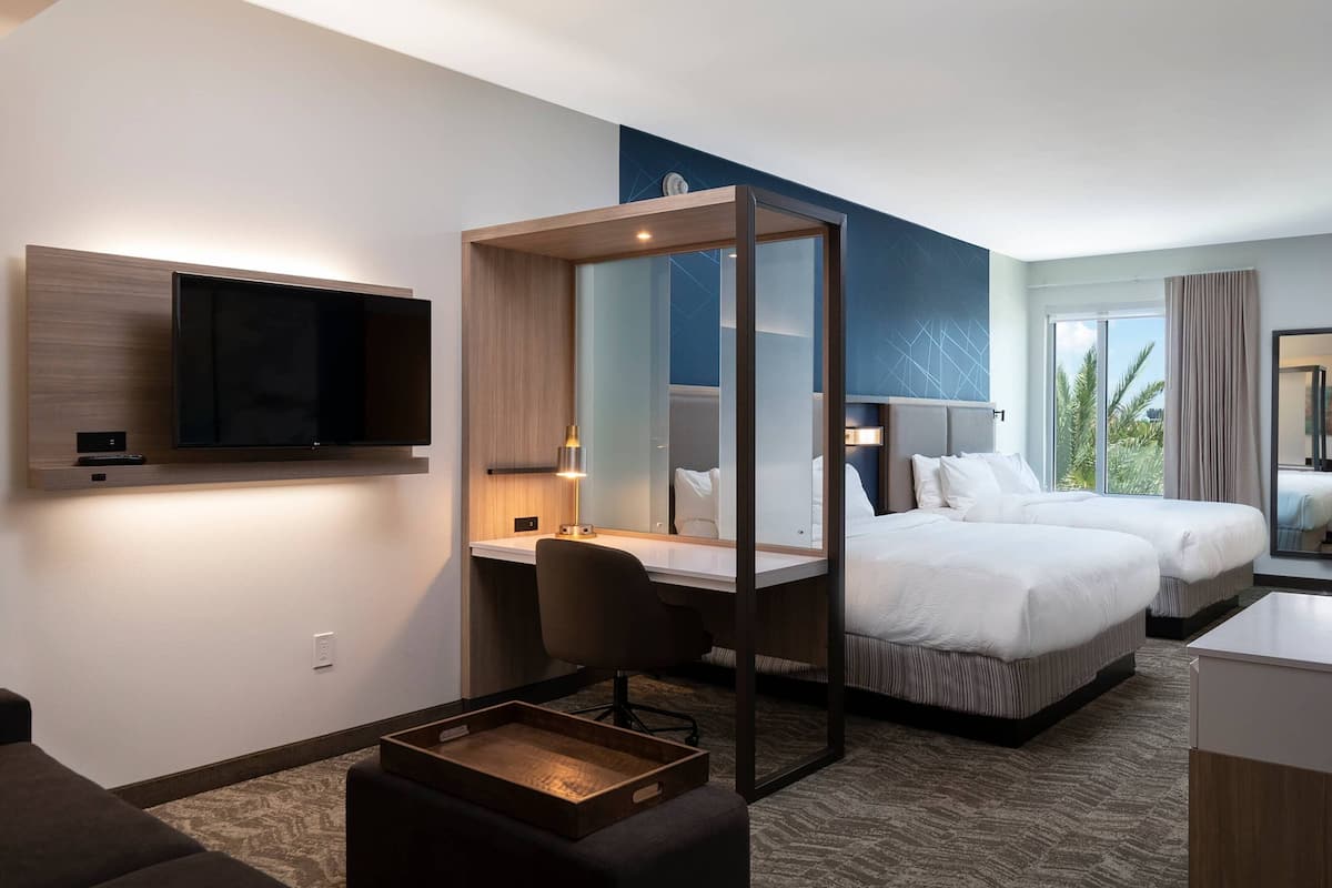 Quarto de hotel com televisão de tela plana grande na parede, uma escrivaninha com cadeira, duas camas, uma pequena mesa de madeira e uma janela grande com cortinas.