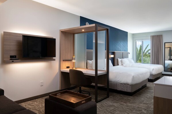 A hotel room with a large flat-screen TV mounted on the wall, a desk with a chair, two beds, a small wooden table, and a large window with curtains.