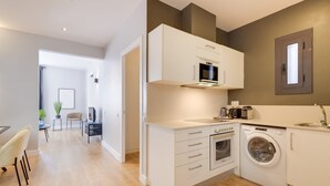 A modern kitchen with a microwave, oven, and washing machine. There is a dining area with a table and chairs visible in the background.