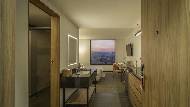 A modern bathroom with a dual sink vanity, a large mirror, a toilet, and a shower area. There is a view of a cityscape through a large window. -  Stadía Suites Querétaro Centro Histórico (Querétaro)