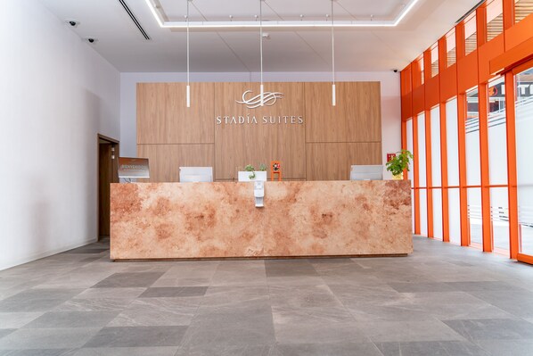 A reception desk with a wooden panel backdrop and a logo, a potted plant, and a view through large windows. - Stadía Suites Querétaro Centro Histórico (Querétaro)