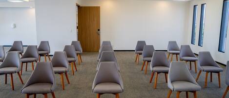 A conference room with rows of grey chairs and wooden legs, carpeted floor, white walls, and a wooden door.