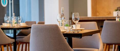 A modern dining area with tables set for two, featuring wine glasses, water glasses, and a small plant centerpiece.