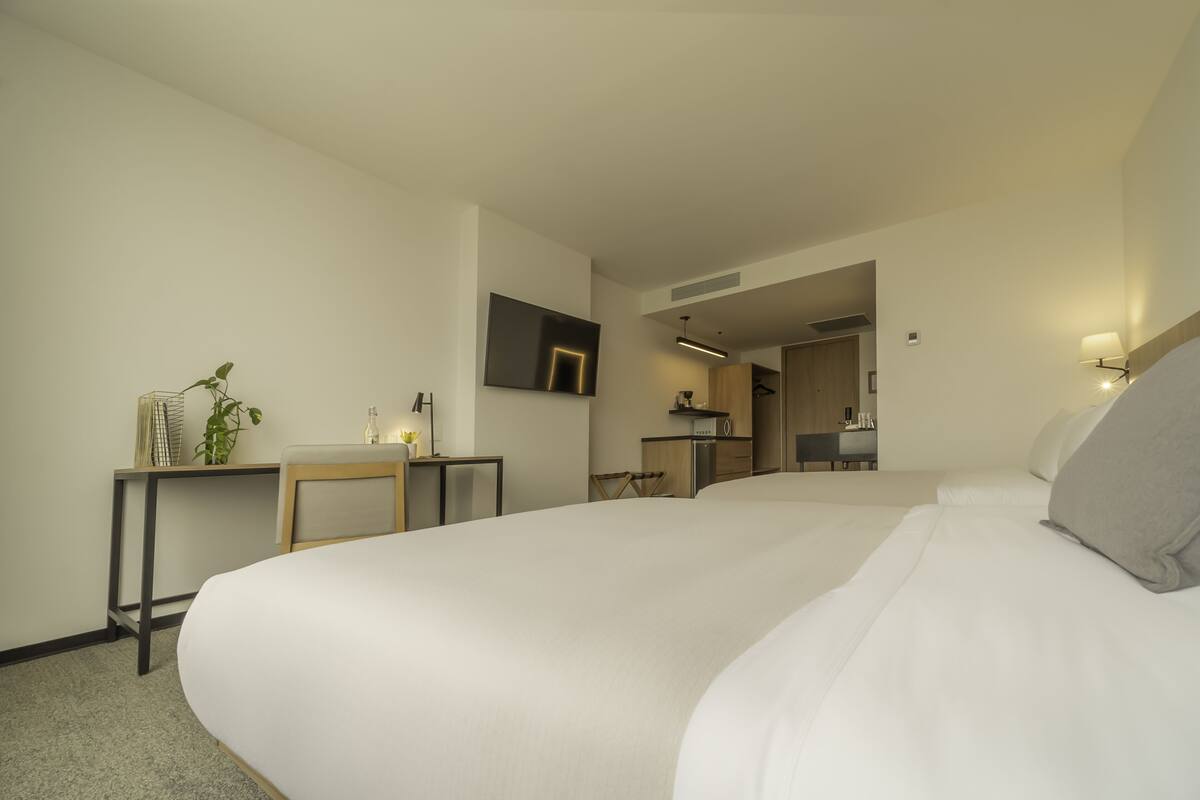 A hotel room with a large bed, a desk, a chair, a small table with a plant, a mirror, and a kitchenette in the background.