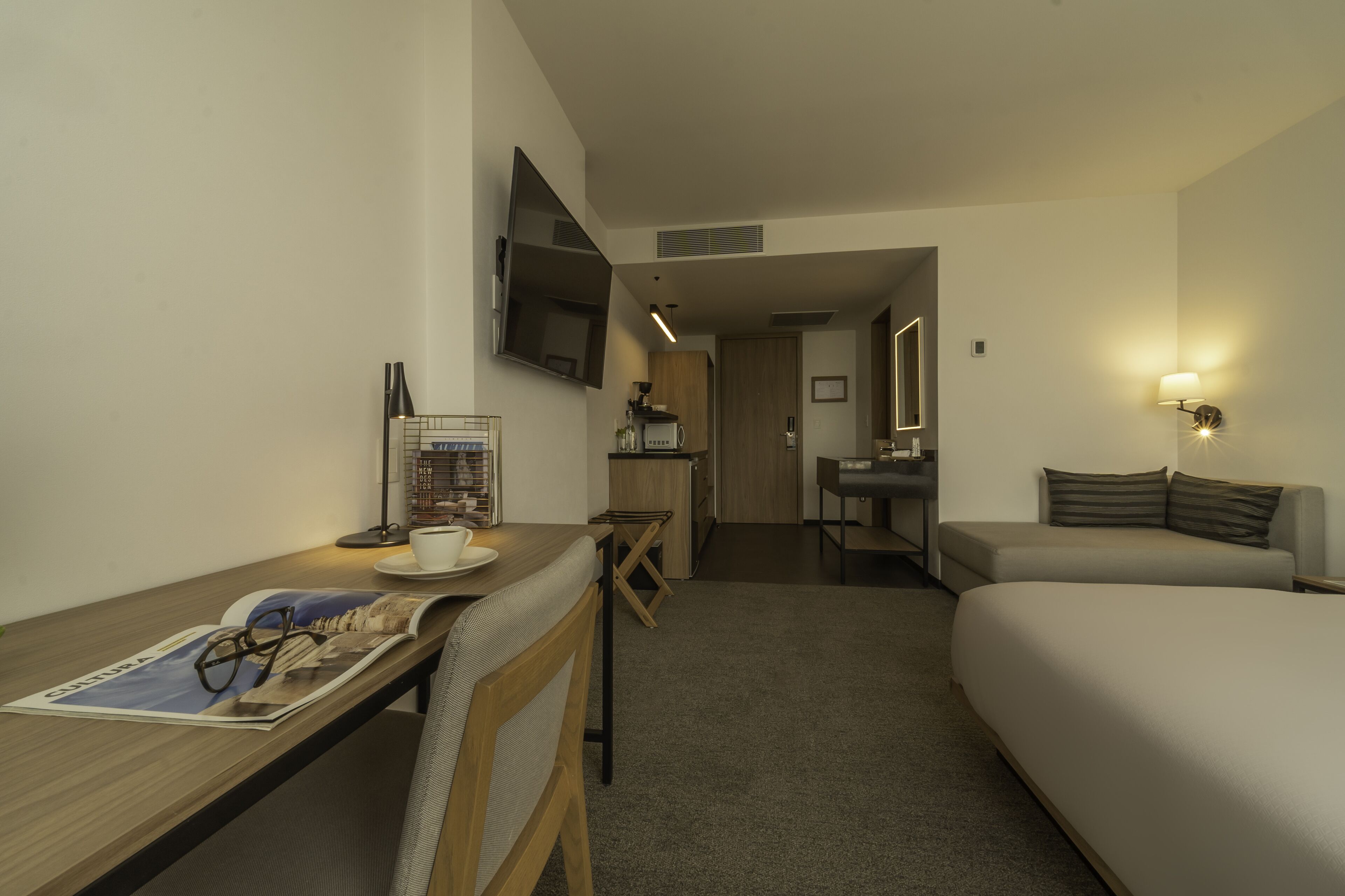 A hotel room with a bed, a desk with a newspaper, a cup, and a pair of glasses.