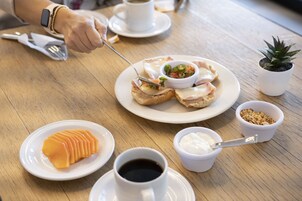 Un desayuno con una fuente de cruasanes, un tazón de fruta, una taza de café y una pequeña planta en maceta sobre una mesa de madera.
