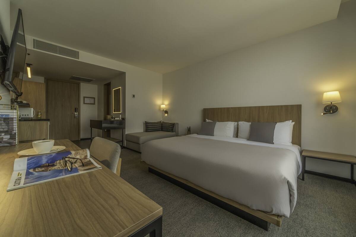 A hotel room with a large bed, a desk with a chair, a television, and a kitchenette area.