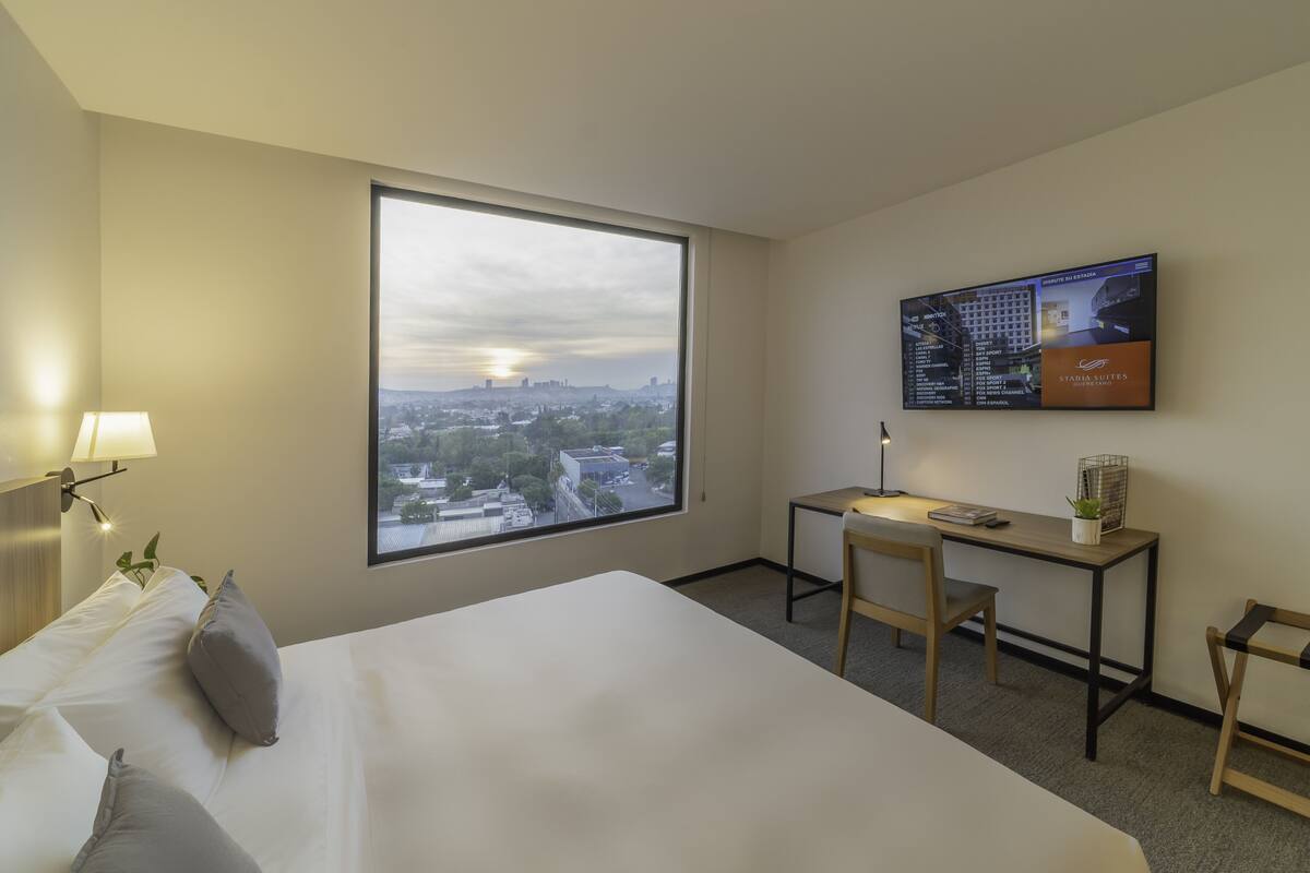 A hotel room with a large window, a bed, a desk, a chair, and a TV displaying a cityscape.