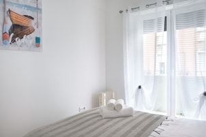 Habitación de hotel con cama, ventana con cortinas traslúcidas, radiador y una obra de arte colgada en la pared.