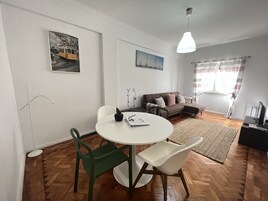 Una sala de estar moderna con una mesa de comedor redonda, sillas verdes y blancas, un sofá y un ventanal con cortinas.