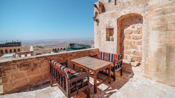 A stone building with a wooden table and benches, a view of the landscape, and a stone wall. - Maristan Tarihi Konak (Mardin)