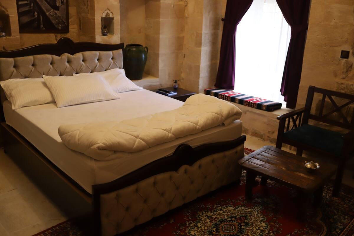 Une chambre à coucher avec un grand lit, une table de chevet en bois, une chaise et une fenêtre avec des rideaux.