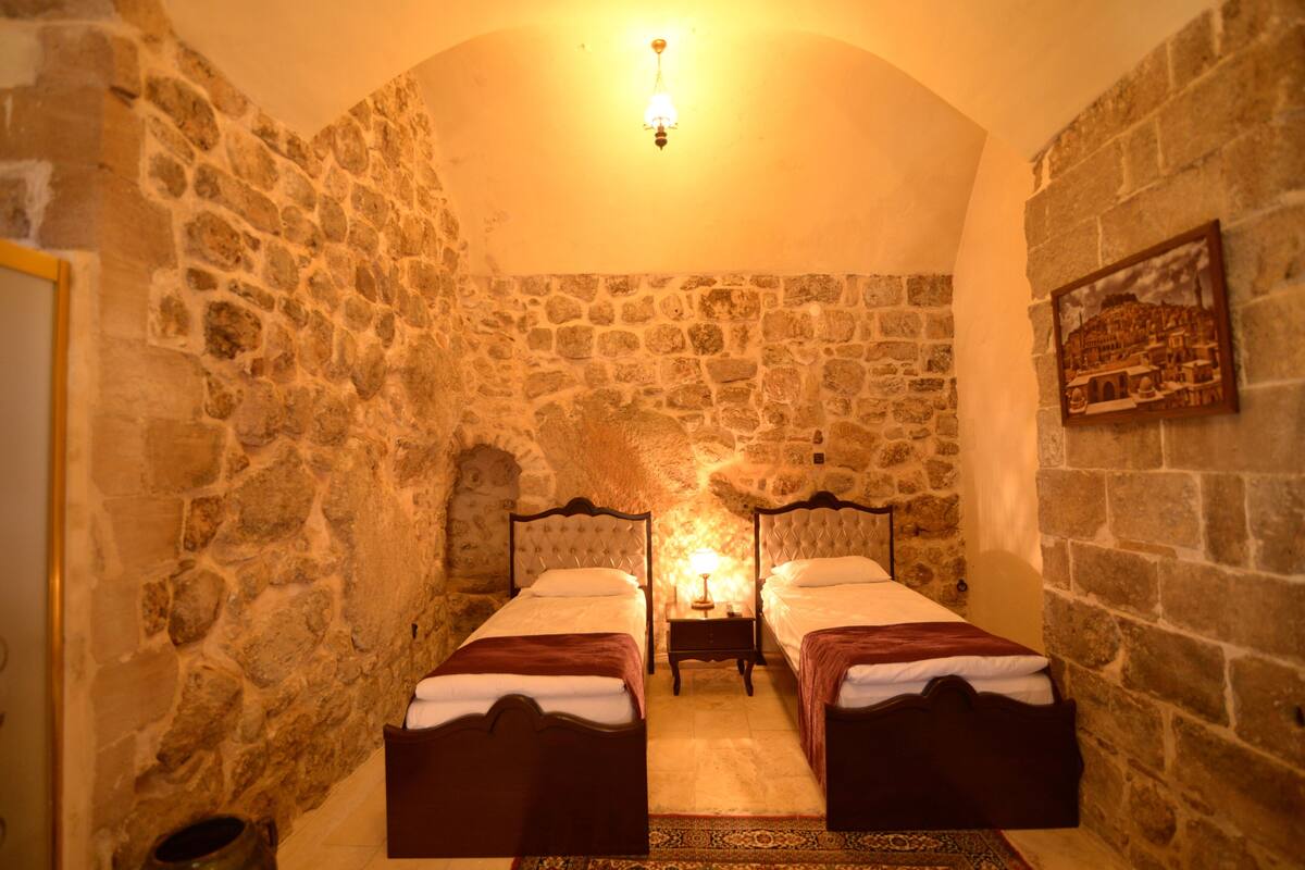 Une chambre avec deux lits, des murs en pierre, une petite table avec une lampe et un tableau encadré au mur.