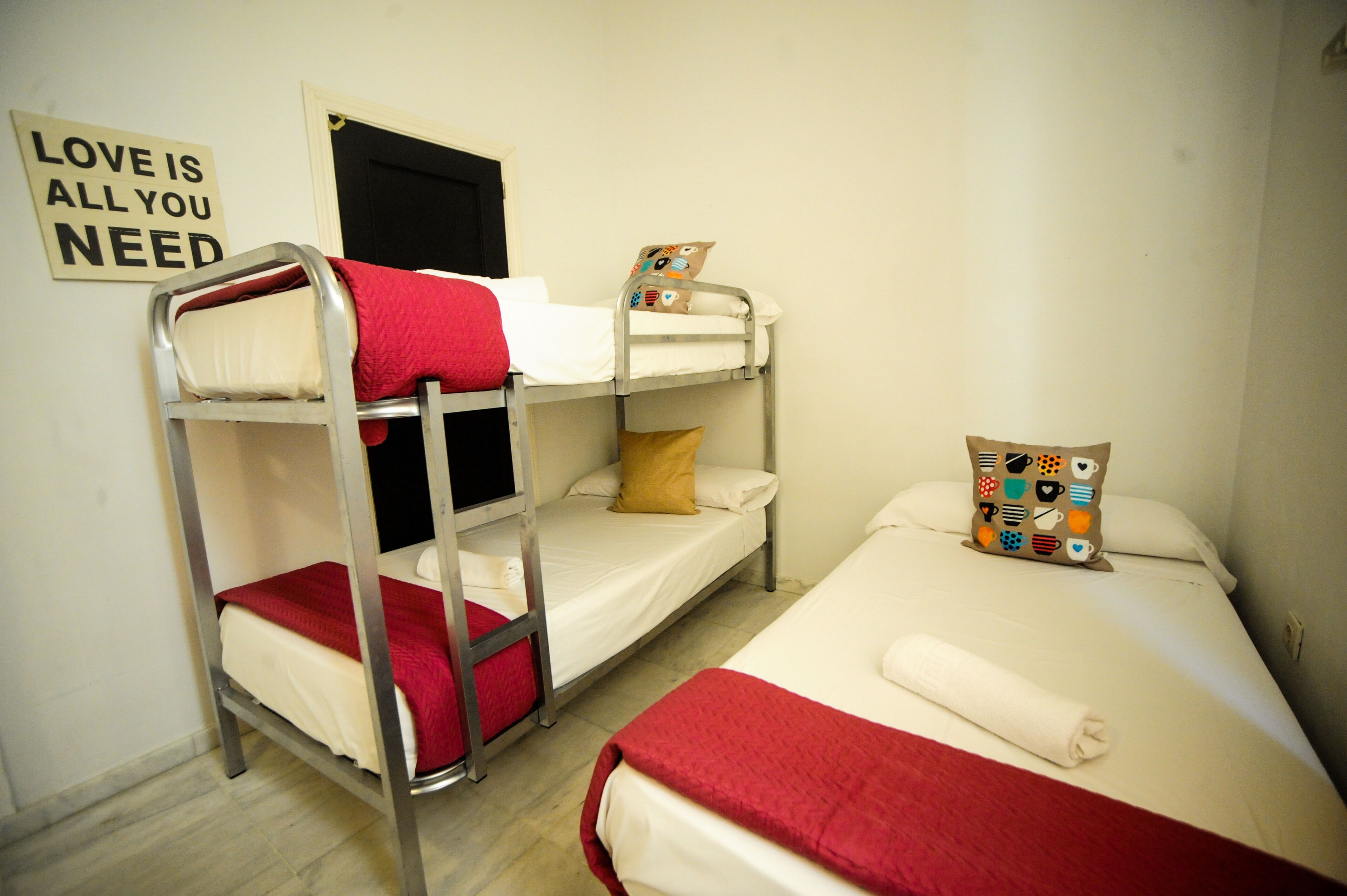 A dormitory room with bunk beds and a single bed, each with pillows and blankets. A sign on the wall reads "LOVE IS ALL YOU NEED."