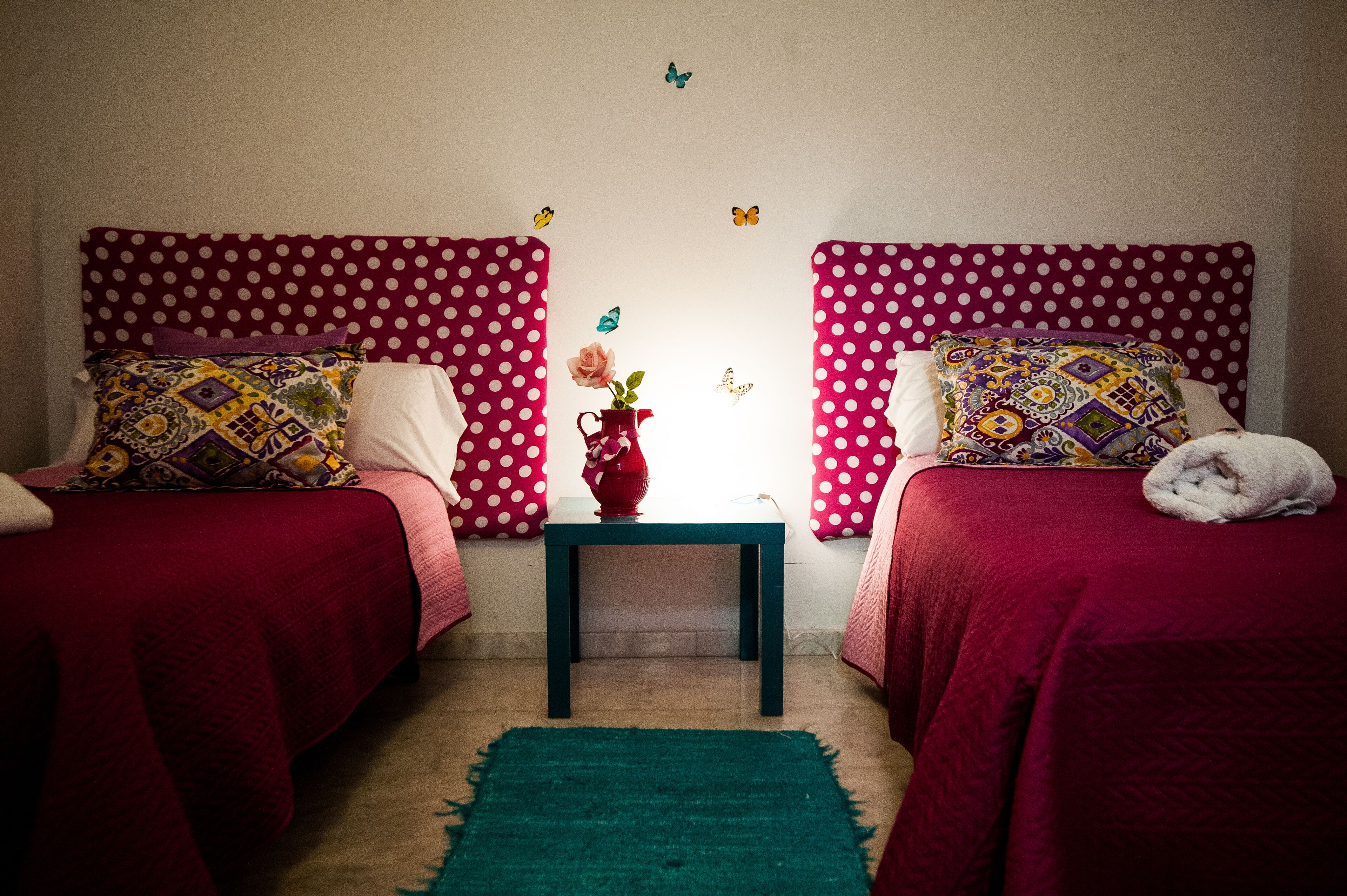 Une chambre avec deux lits, chacun doté d’une tête de lit à motifs à pois et de draps rouges. On y trouve une petite table sur laquelle est posé un vase de fleurs, ainsi que des autocollants décoratifs représentant des papillons sur les murs.