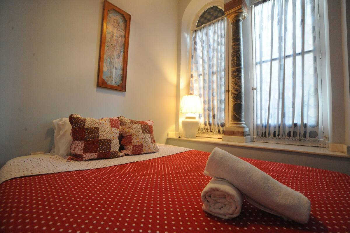 Siististi pedattu sänky, jossa on punavalkoinen pilkkukuviollinen päiväpeite, kaksi tyynyä ja rullalle taitettu pyyhe sängyn vieressä.
