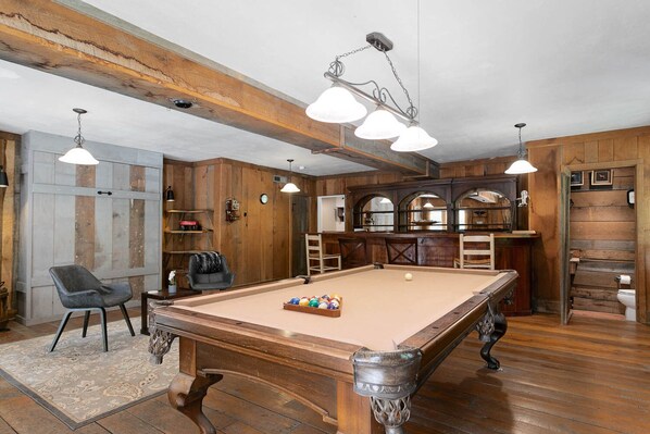 A pool table in a room with wooden walls and ceiling beams, a bar area with stools, and a clock on the wall.
