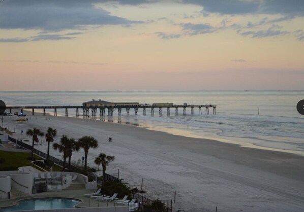 A beach with a pier extending into the ocean, a pool area, and palm trees.