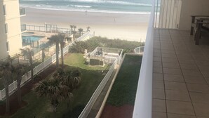 A balcony with a view of the beach, a pool, and a grassy area with palm trees. - Luxury Ocean View Condo with Access to No Drive Beach (Daytona Beach Shores)