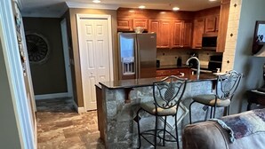 A kitchen with a central island, wooden cabinets, and a refrigerator. There is a bar stool and a brown leather couch in the foreground. - Luxury Ocean View Condo with Access to No Drive Beach (Daytona Beach Shores)