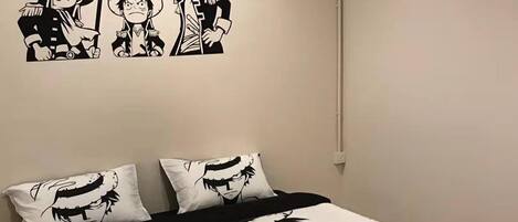 Un dormitorio con una cama que presenta un estampado de gran tamaño al estilo manga, almohadas con diseños similares y una decoración mural sobre la cama con personajes de manga.