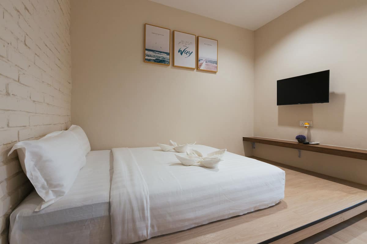 Una cama bien hecha con sábanas blancas y almohadas decorativas, un televisor de pantalla plana montado en la pared y tres cuadros enmarcados sobre la cama.